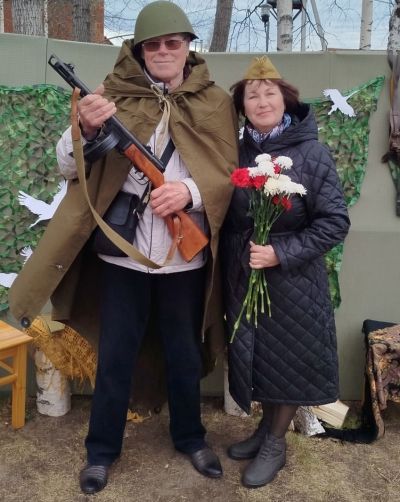 мужчина в плаш-палатке и каске с автоматом и женщина в пилотке с гвоздиками