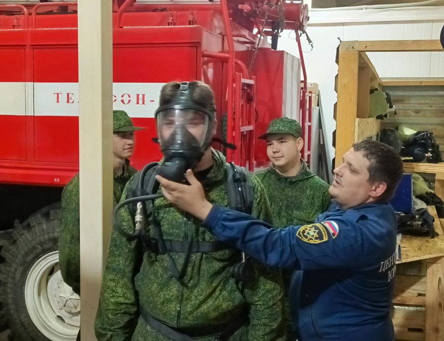 Сотрудник помогает восьмикласснику примерить боевое снаряжение пожарного